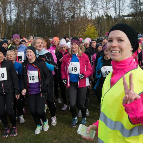 löpare står startklara för att springa ett lopp. De har nummerlappar på magen och en funktionär i väst ler och gör segertecken.