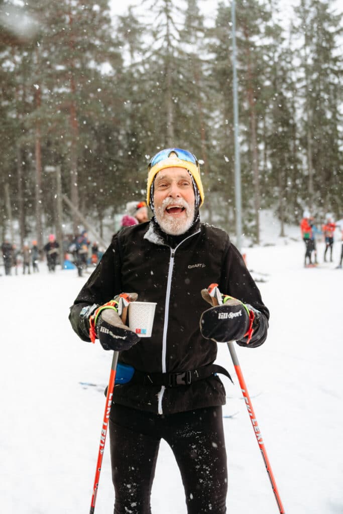 En man med skidor och skidstavar står i snön med en kopp blåbärssoppa i handen.