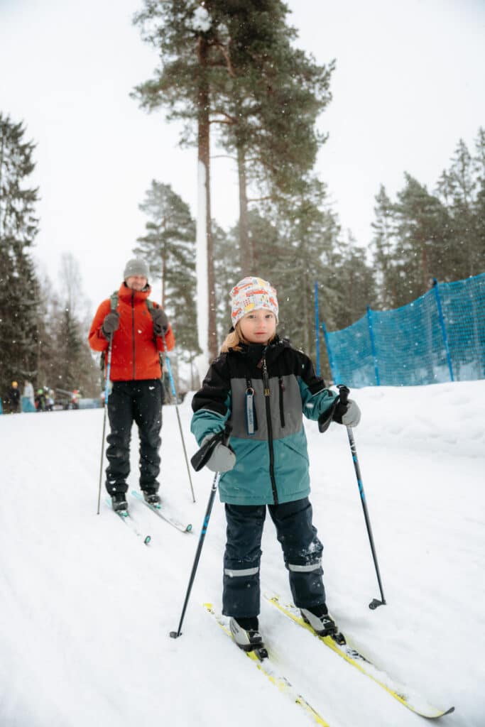 En barn åker längdskidor på snön med sin pappa efter sig.