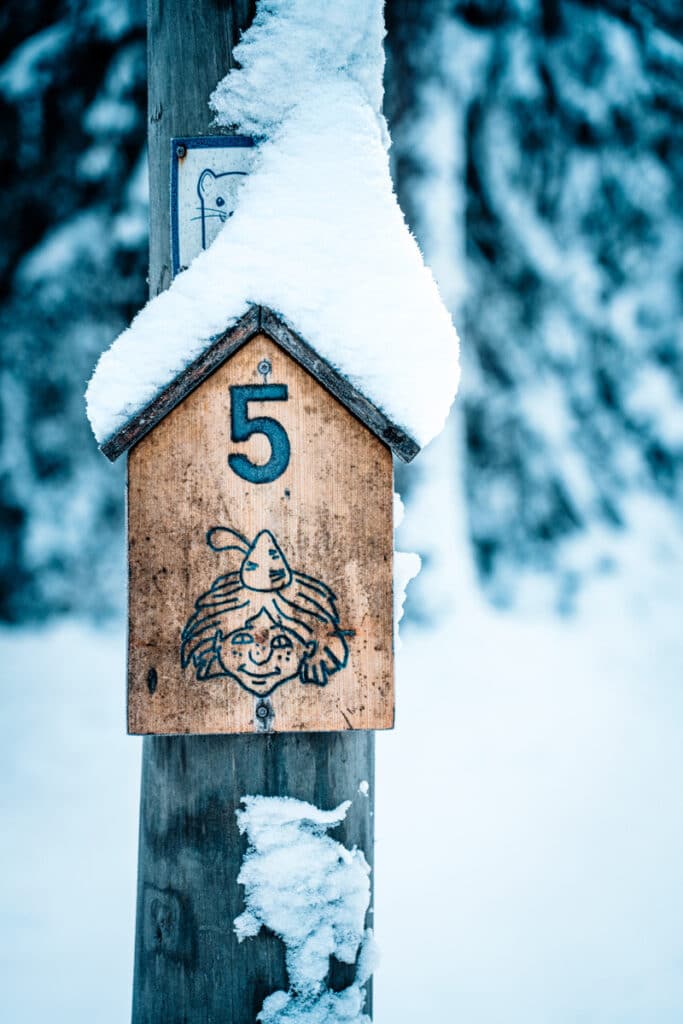 En skylt i trä med Skogsmulle och nummer 5. Skylten är fast i ett träd och har snö på sig.