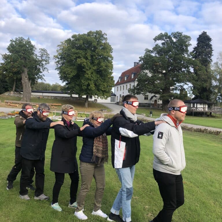 Sex personer står utomhus på rad med händerna på varandras axlar, med ögonbindel.