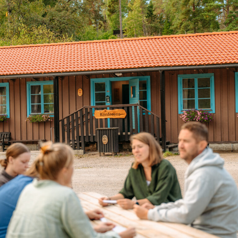 En grupp människor som sitter runt ett picknickbord. I bakgrunden syns en konferenslokal.